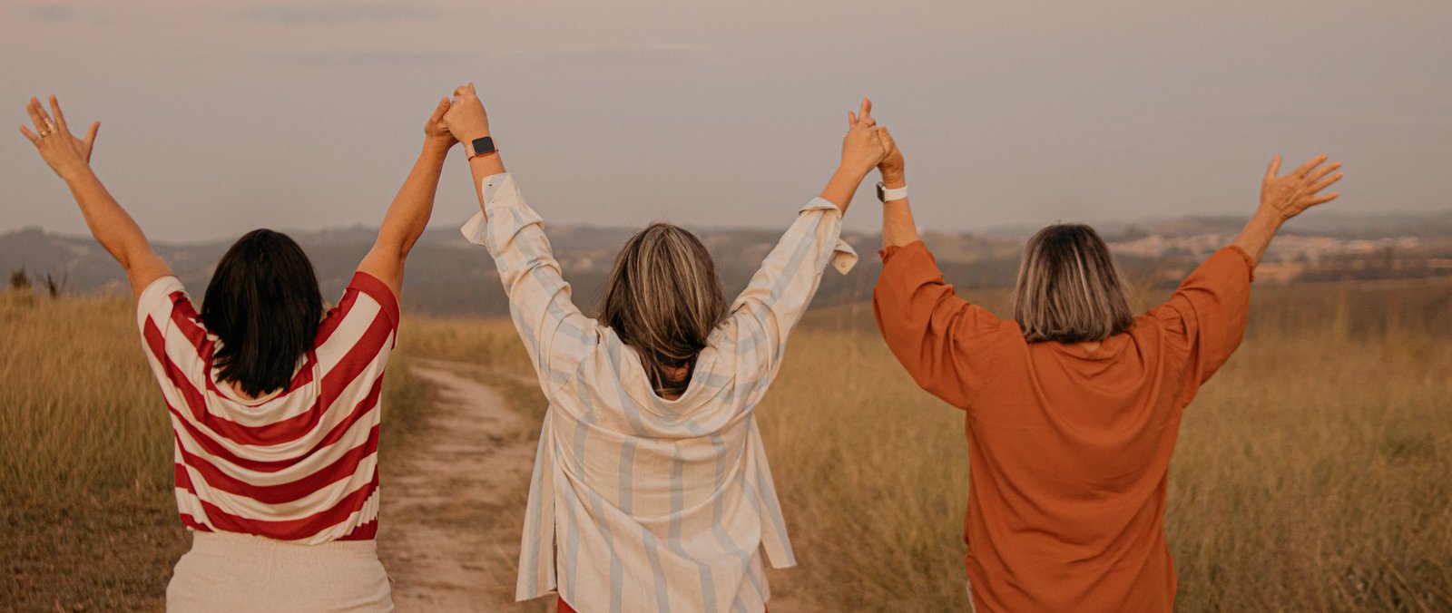 Three women stand together in a golden-lit field, arms raised and hands joined, facing an open path ahead